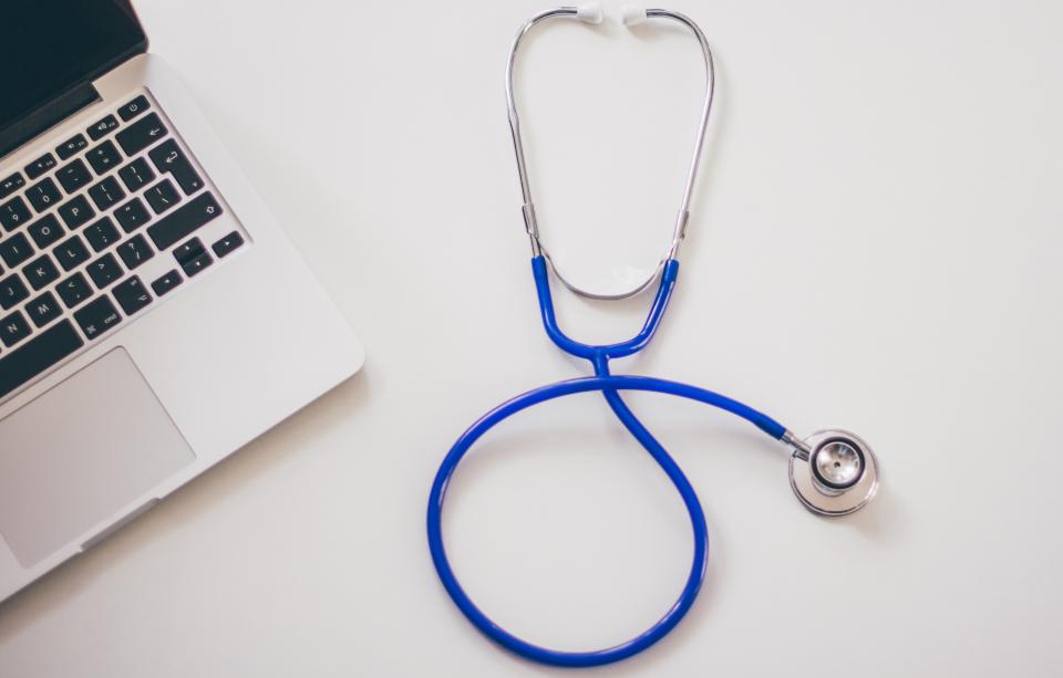 A stethoscope resting beside an open laptop on a clinic desk